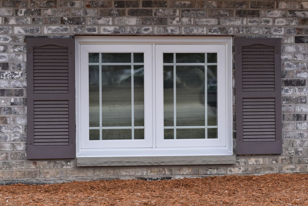 A white window with brown shutters on a brick wall