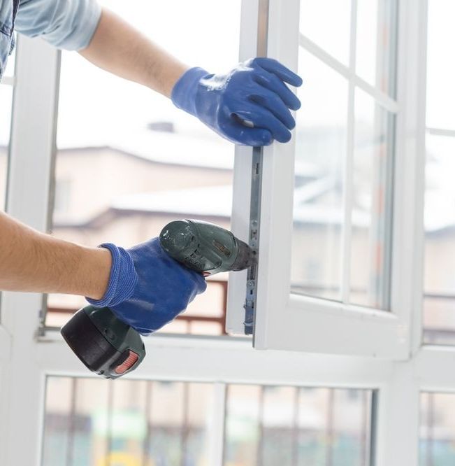 Person in blue gloves using a power drill to install a white window.