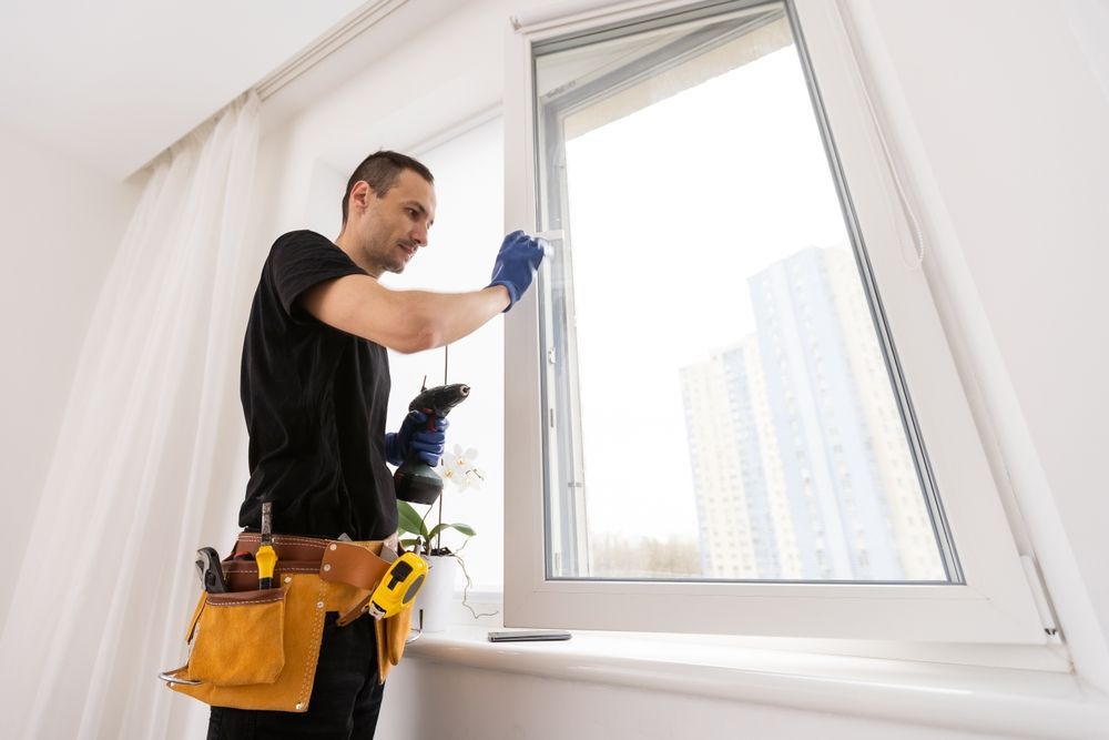 Man with tool belt installs window in a white-walled room with a city view.