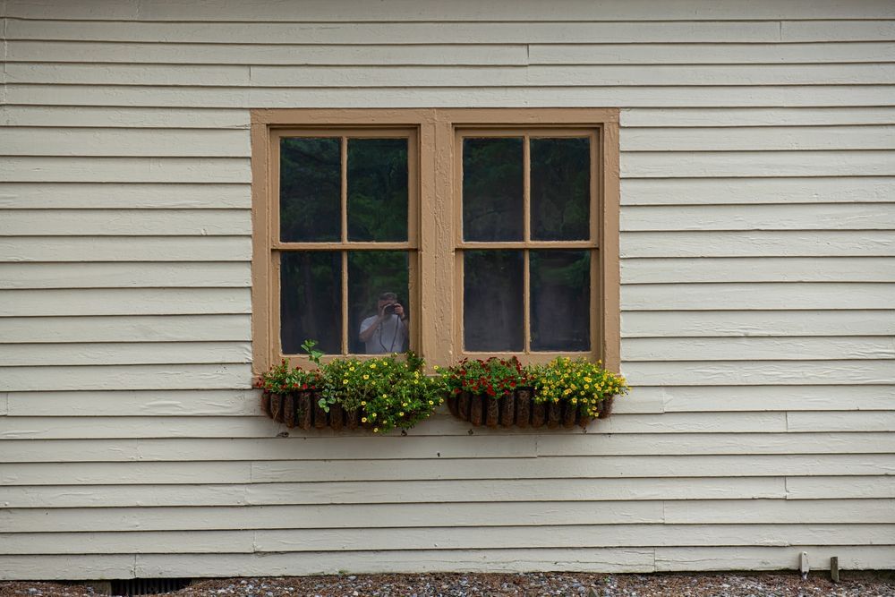 There are two windows on the side of a house with flowers in them.
