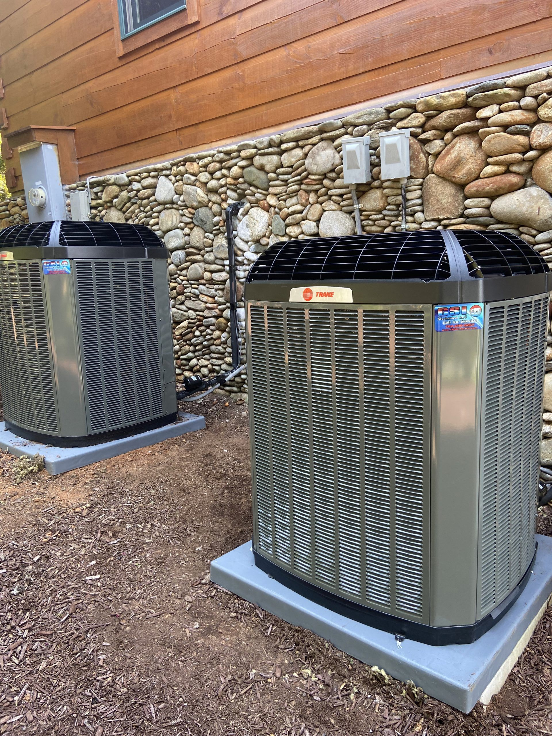 Two Trane air conditioning units sitting on gray concrete pads against a stone wall and wooden siding.