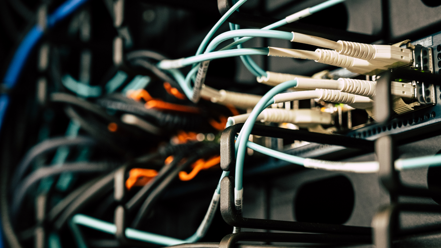 Close-up of tangled network cables plugged into a server rack, with blue, white, and orange wires.