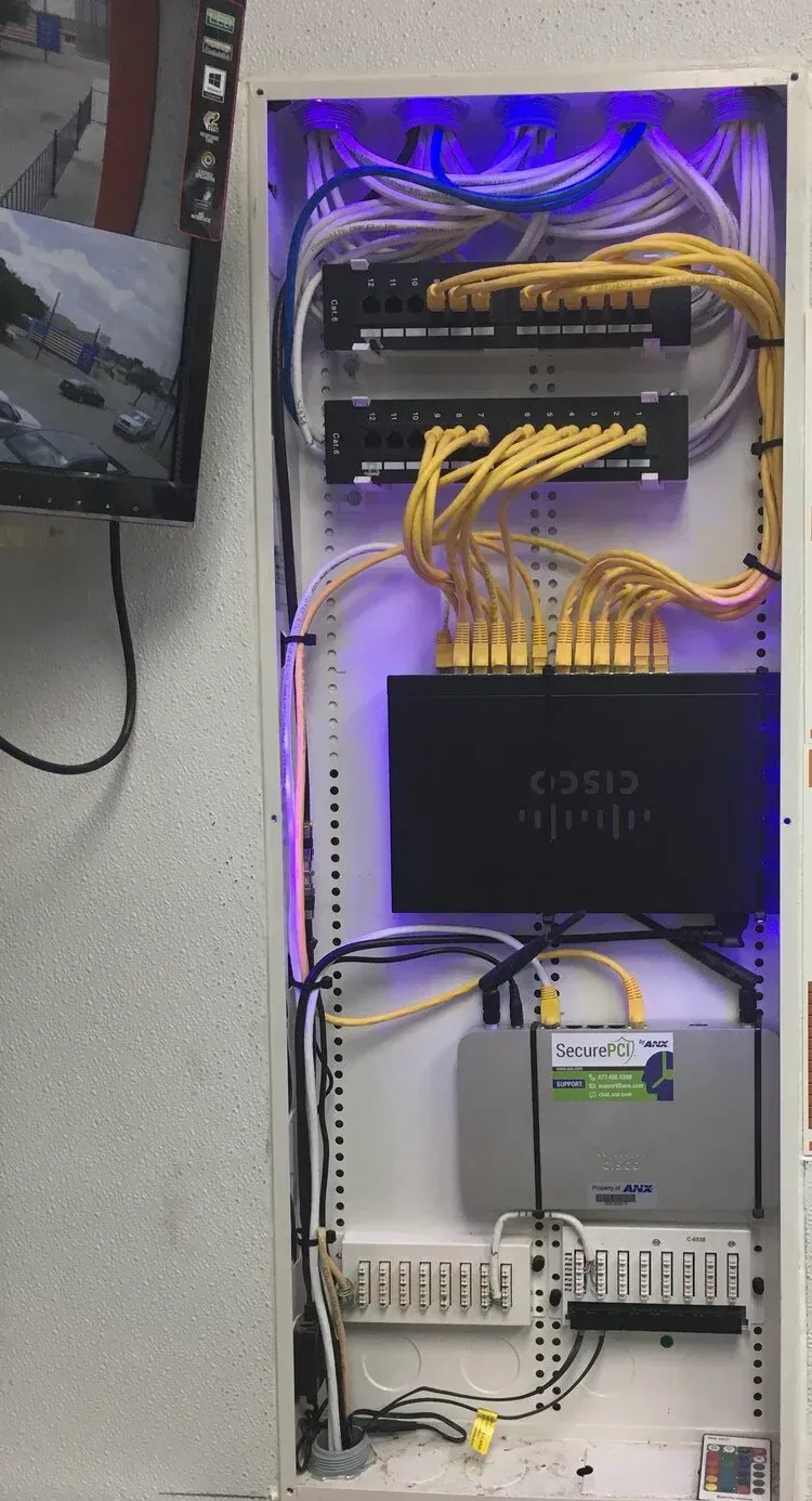 Wall-mounted network rack with Ethernet switches, patch cables, and a modem under purple lighting