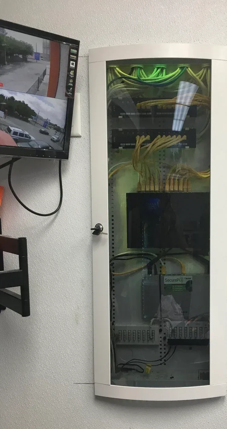 Wall-mounted glass server cabinet with cables and equipment, next to a monitor on a white wall.