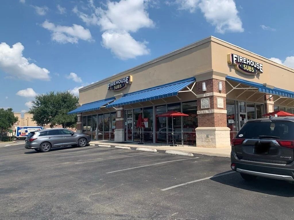 Strip mall storefronts with blue awnings, parked cars, and a partly cloudy sky
