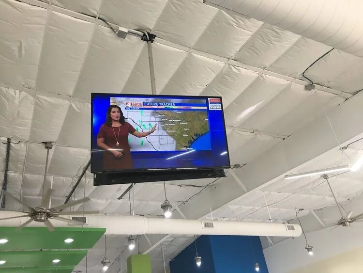 TV screen hanging from a white ceiling in a large indoor space, showing a weather broadcast.