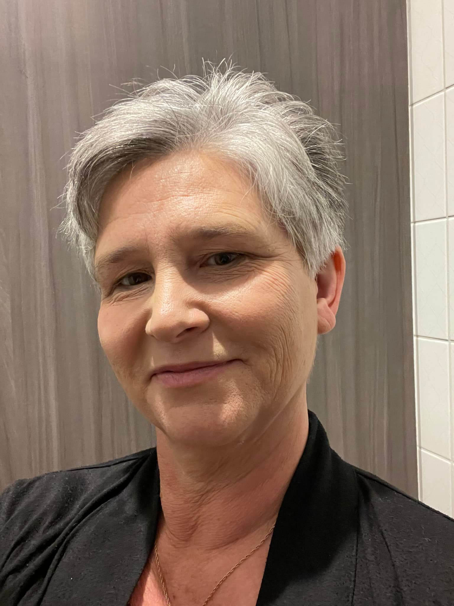 A person with short, gray hair and a neutral expression, wearing a black shirt against a gray wall and white tile background.