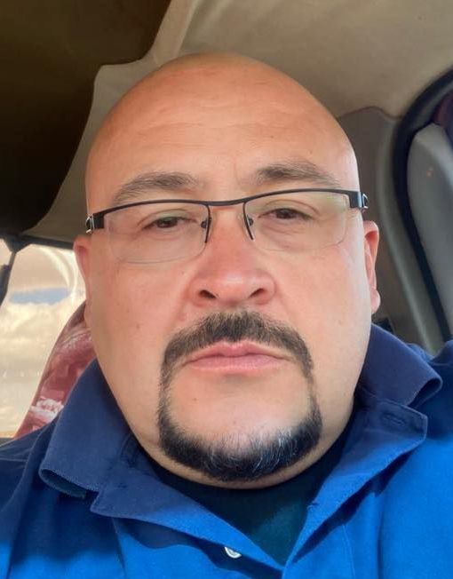 A person with a goatee and glasses wearing a blue collared shirt, shown in a headshot taken inside a vehicle.