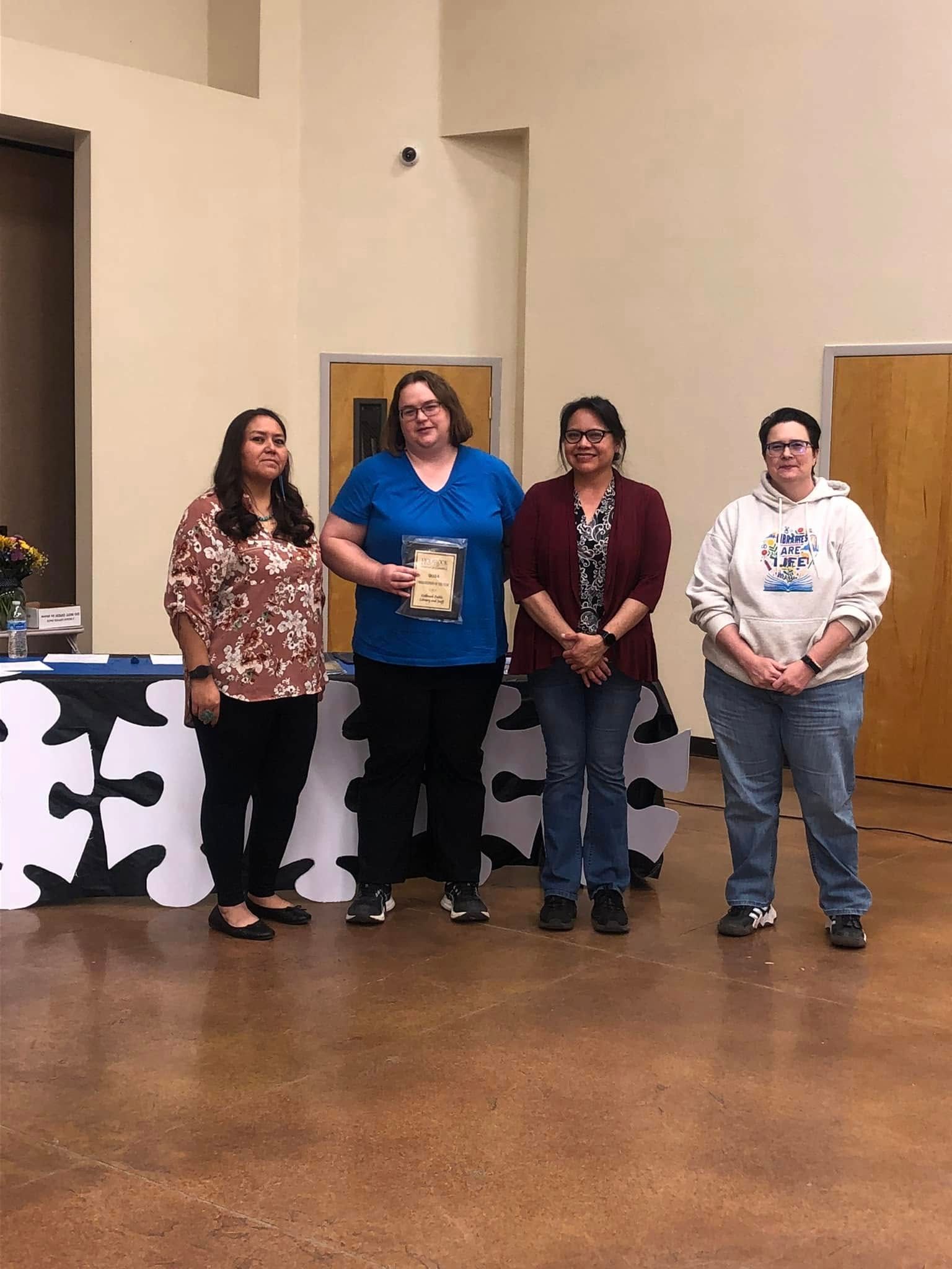Four people stand in a line indoors, with one person in the center holding an award plaque in front of a decorative table.