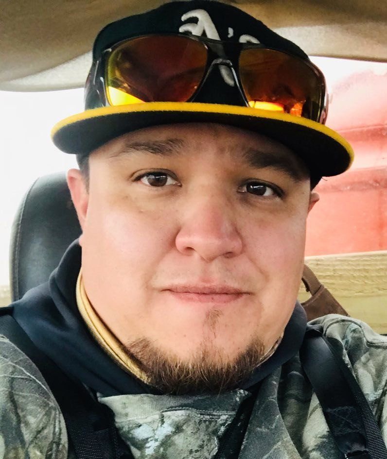 A person wearing a black and gold baseball cap and amber-tinted sunglasses, dressed in camouflage clothing.