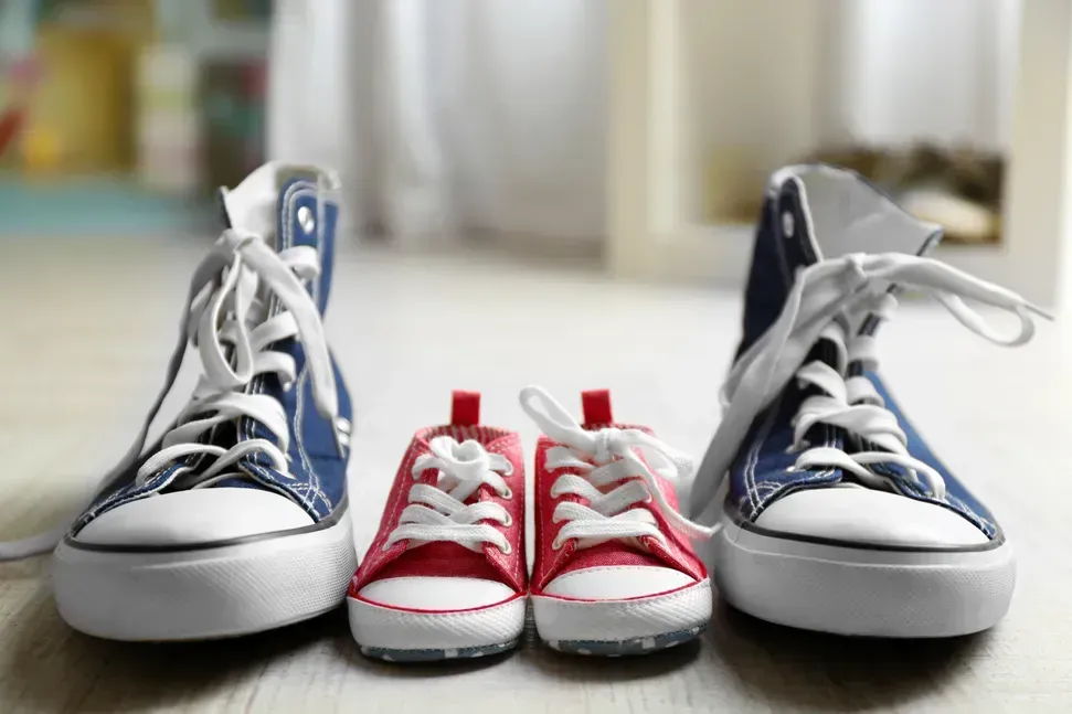 Blue adult sneakers flank red baby sneakers on a wooden floor indoors.