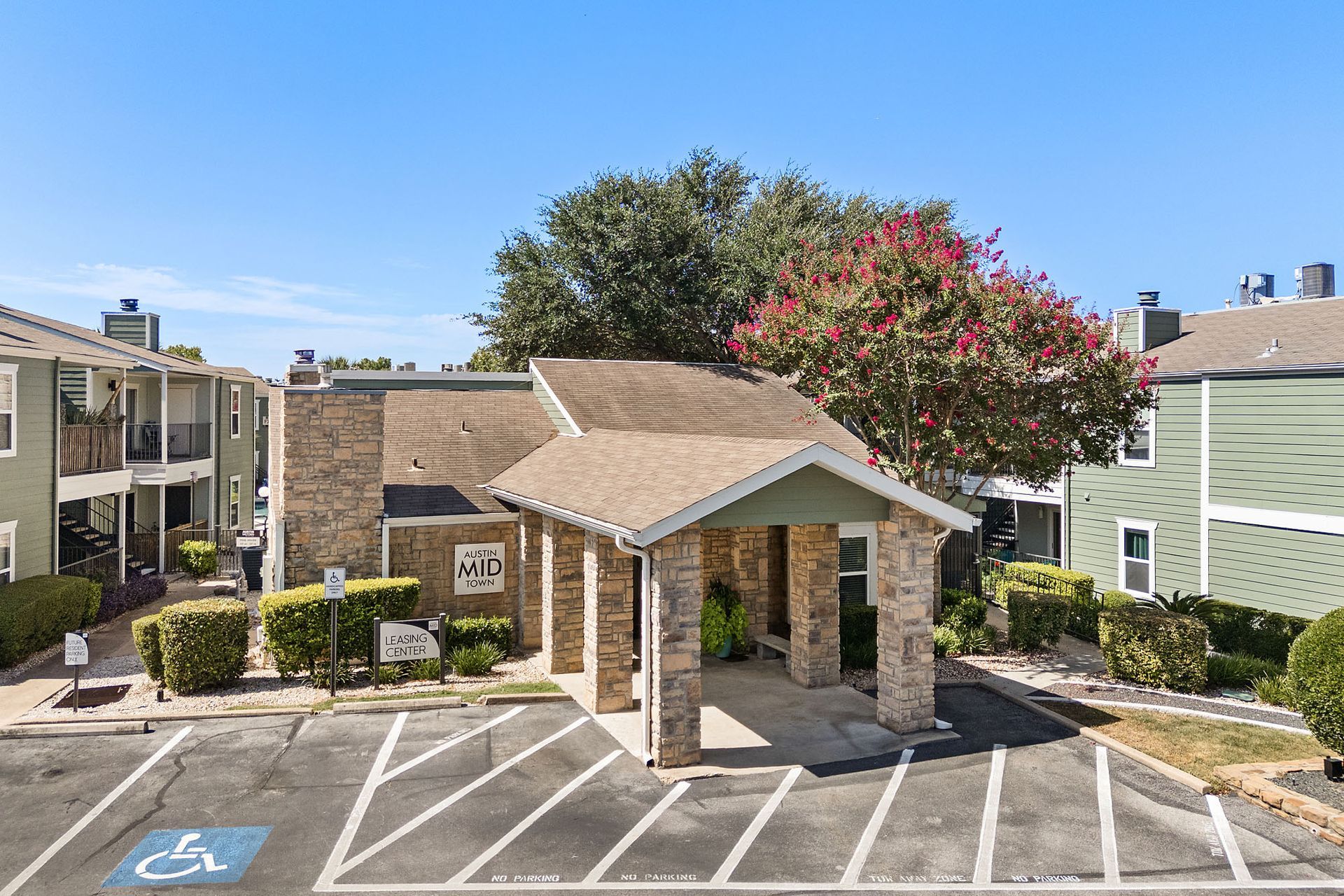 Exterior view of Austin Midtown apartment complex's leasing center with a stone facade entrance and covered walkway, surrounded by parking spaces and nestled between residential buildings under a clear blue sky at Austin Midtown in North Austin, TX.