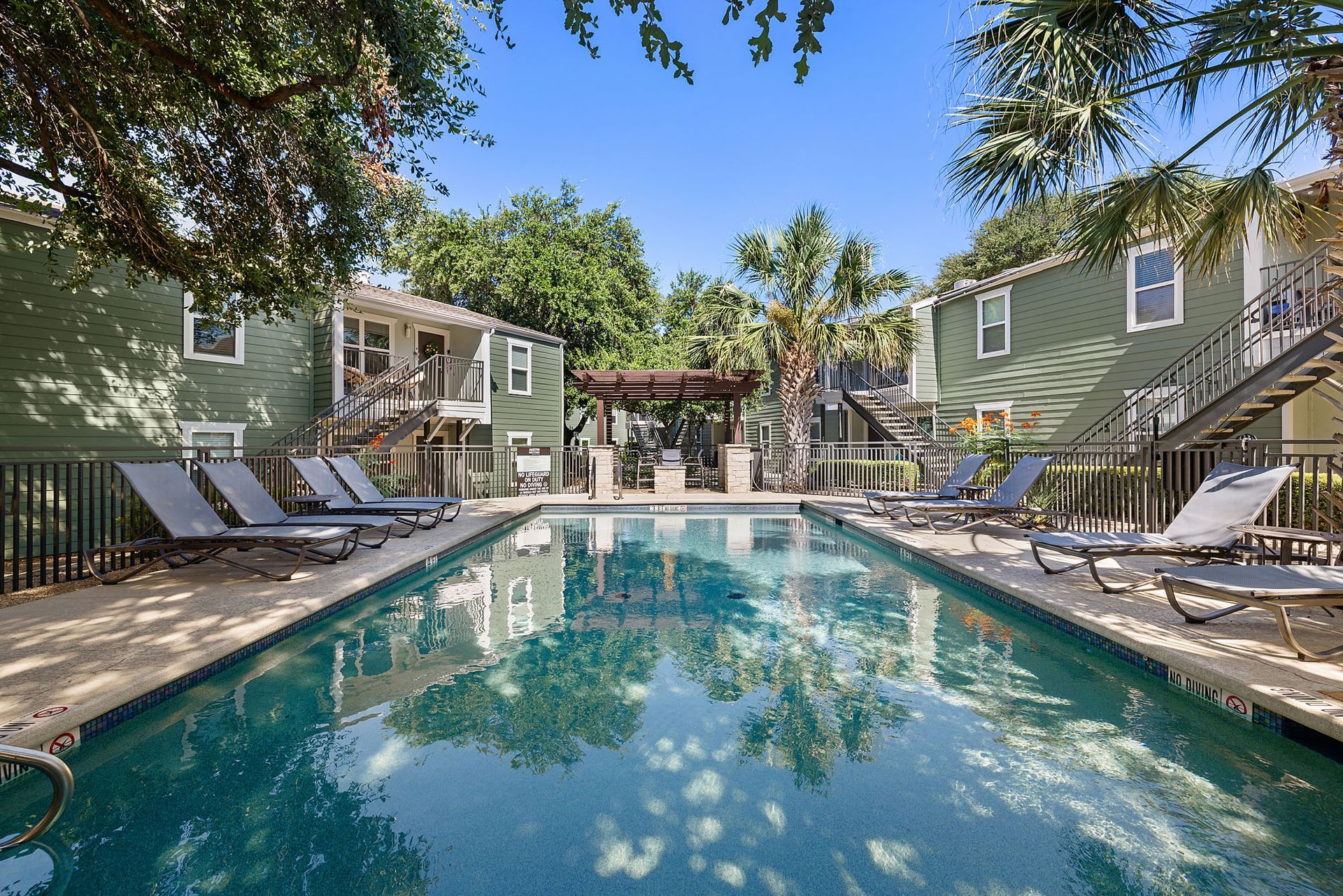 Apartment complex pool with lounge chairs, trees, and turquoise water in a courtyard