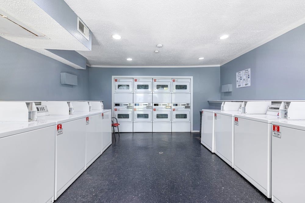 Interior of a well-lit, clean laundry facility with multiple front-loading washing machines on the left and stacked dryers on the right. The walls are painted blue and grey, and there's a speckled grey floor. Instructions and signs are visible on the wall at Austin Midtown in North Austin, TX.