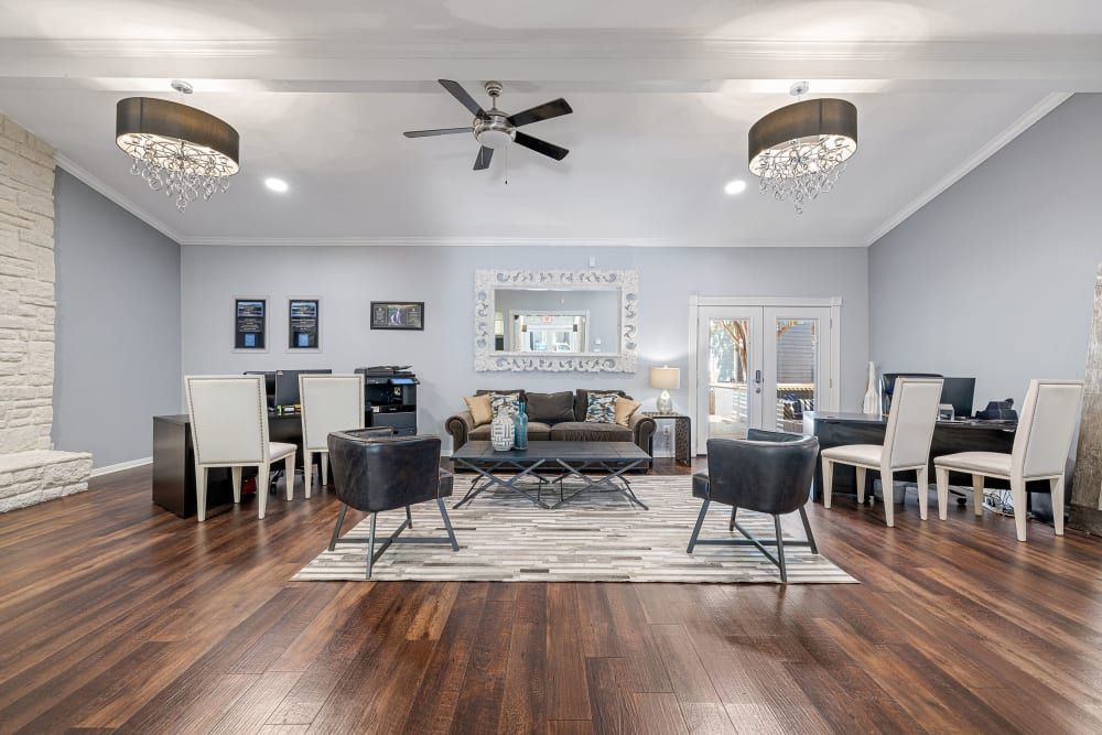 Modern clubhouse with hardwood floors and a neutral color palette featuring a seating area with a dark blue sofa and armchairs, a stone accent wall, and an office space with a desk and chair. The dining area includes a white table with matching chairs. Elegant light fixtures hang from the ceiling, and natural light enters through glass doors at Austin Midtown in North Austin, TX.