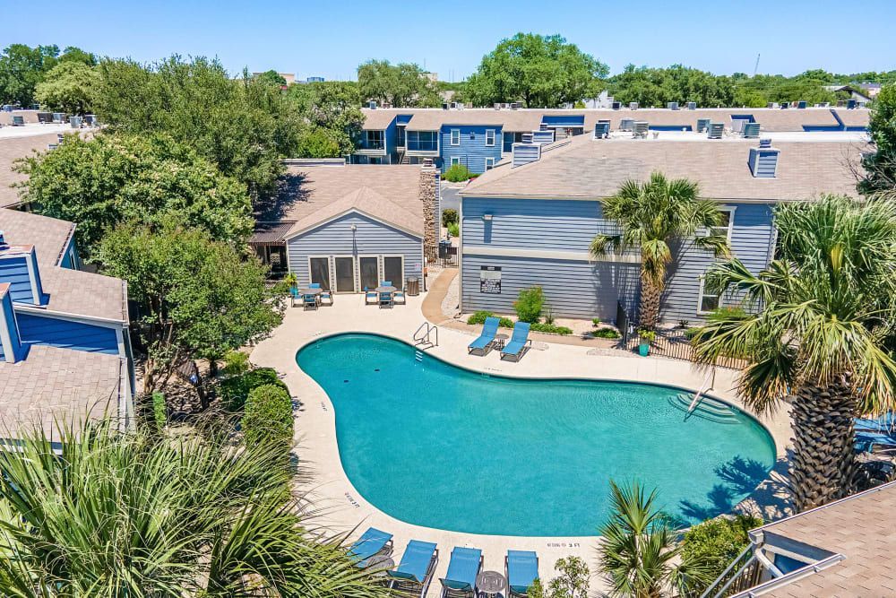 Aerial view of a residential complex featuring light gray buildings with blue trim and gabled roofs. In the center, there is a kidney-shaped swimming pool surrounded by sun loungers, with palm trees and greenery adding a lush aspect to the setting at Austin Midtown in North Austin, TX.