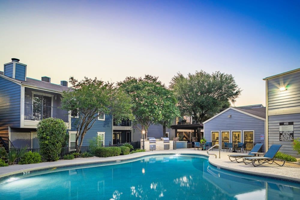 Apartment courtyard with a turquoise pool, lounge chairs, and lit buildings at dusk at Austin Midtown offers apartments in North Austin, TX.