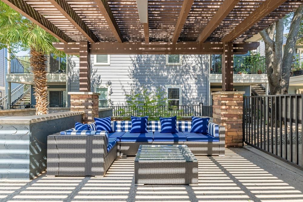 Outdoor patio area with a pergola casting shadow patterns on the ground, featuring modern wicker furniture with blue and white striped cushions, a glass-top coffee table, surrounded by a metal railing, palm tree, and multi-level residential buildings in the background at Austin Midtown in North Austin, TX.