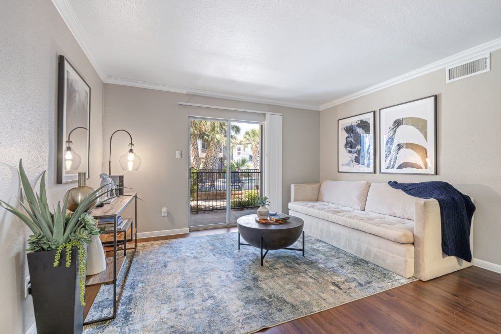Modern living room interior with a beige sofa, decorative throw blanket, and a round coffee table on a blue distressed area rug. The room features wall-mounted light fixtures, abstract wall art, a tall plant in a black vase, and a narrow console table with a mirror. Sunlight streams in through a glass sliding door that opens onto a balcony with a view of palm trees at Austin Midtown in North Austin, TX.