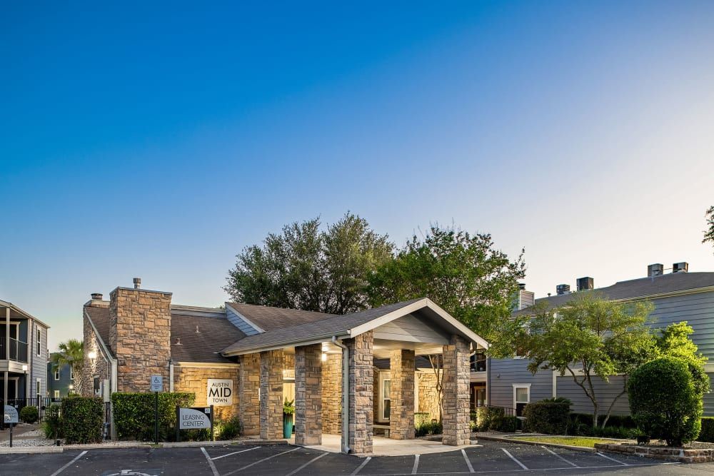 Exterior view of Austin Midtown apartment complex's leasing center with a stone facade entrance and covered walkway, surrounded by parking spaces and nestled between residential buildings under a clear blue sky at Austin Midtown in North Austin, TX.