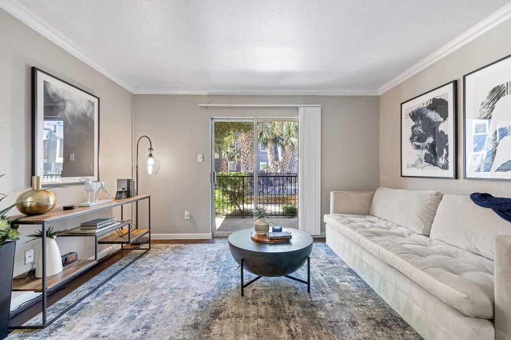 A well-lit contemporary living room featuring a plush beige sofa, a round black coffee table, and a glass console table with decorative items. The room is adorned with abstract wall art and includes a floor lamp. Multicolored area rug anchors the space, while sliding glass doors lead to an outdoor area with greenery at Austin Midtown in North Austin, TX.
