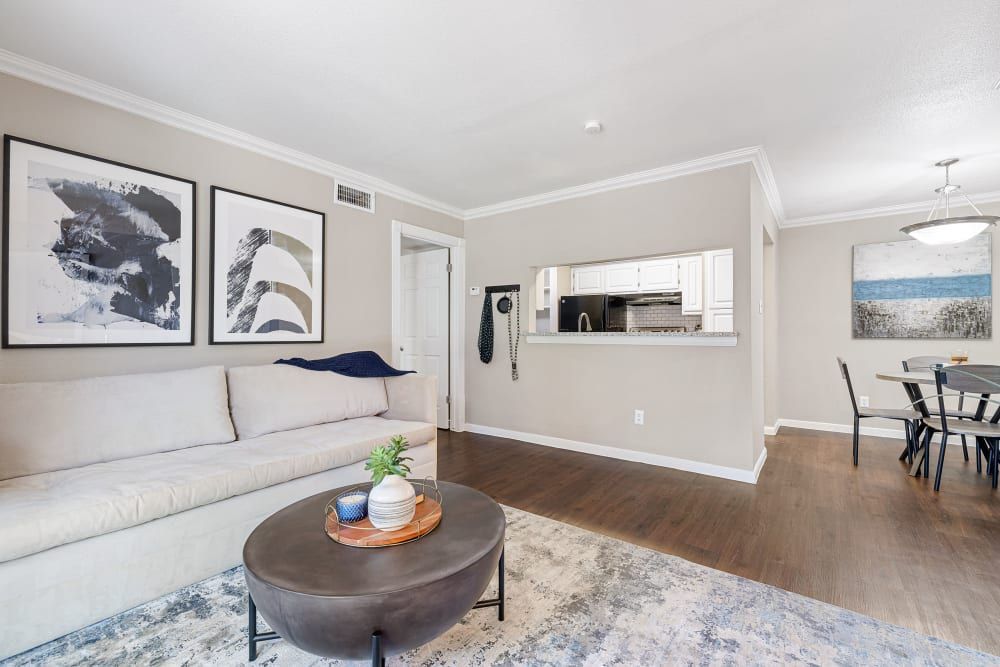 Modern living room seamlessly connected to a dining area, featuring a large beige sofa with blue pillows, abstract wall art, a round coffee table with decorative items, and a view into a kitchen with wooden cabinets and dark countertops at Austin Midtown in North Austin, TX.