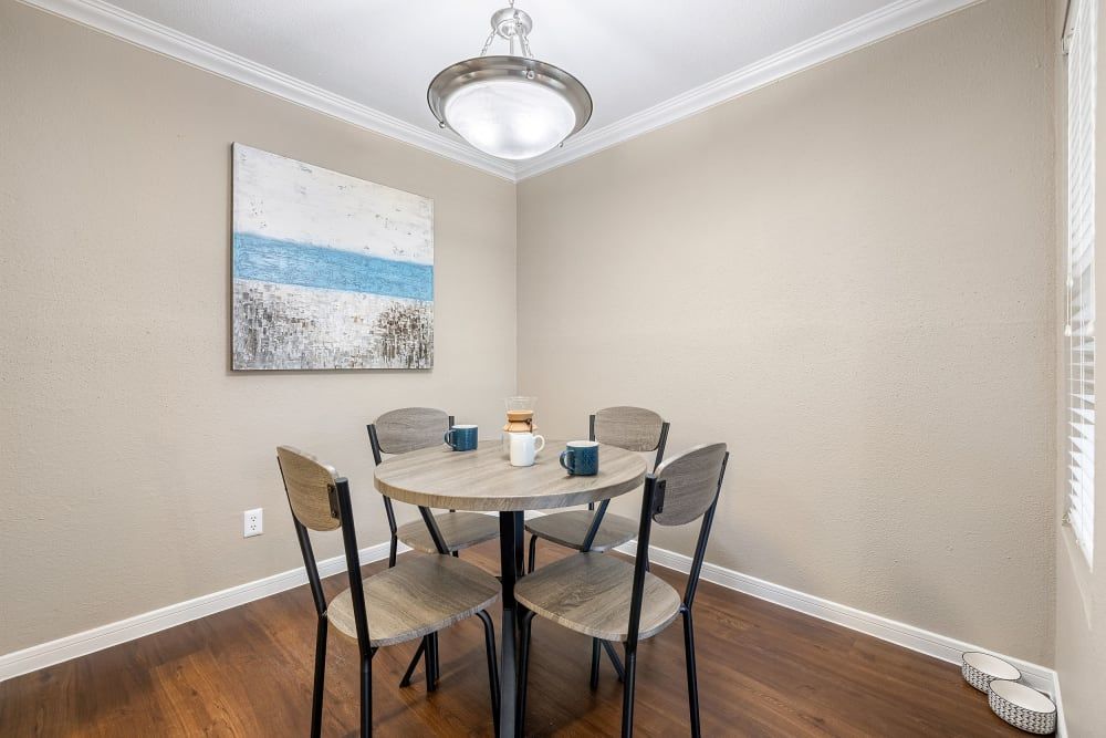 Cozy dining nook with a modern round table set for two, featuring wooden chairs with black metal legs on a dark hardwood floor. An abstract ocean painting adorns the beige wall above, complemented by a simple light fixture. White blinds cover a window on the right, and decorative baskets sit on the floor by the wall at Austin Midtown in North Austin, TX.