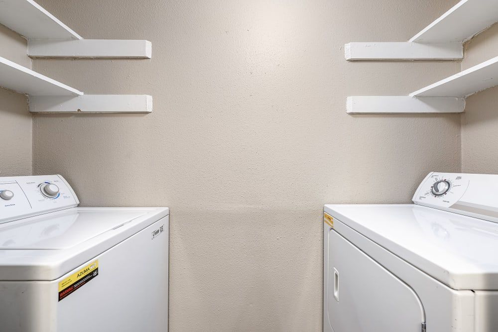 A compact laundry room featuring a top-loading washing machine on the left and a front-loading dryer on the right, with three white empty shelves mounted on the beige wall above them at Austin Midtown in North Austin, TX.