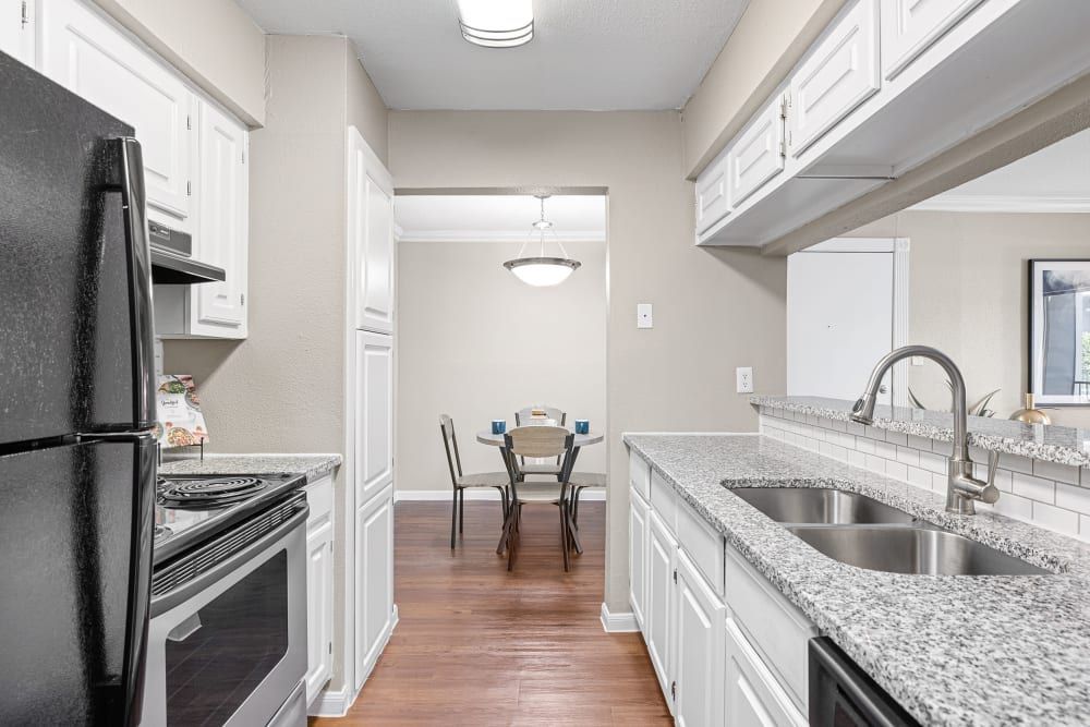 Modern kitchen interior with white cabinetry and granite countertops, featuring stainless steel appliances including a refrigerator, stove, and dual sink. There is a dining area with a wooden table and chairs in the background, set on hardwood flooring. The space is well-lit with natural light from a window and artificial light from a pendant lamp over the dining table at Austin Midtown in North Austin, TX.