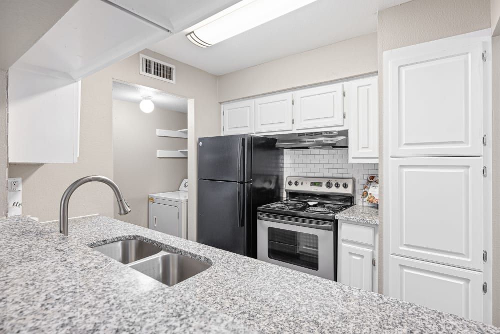 Modern kitchen interior with white cabinets, granite countertops, stainless steel sink, a black refrigerator, and a matching black stove/oven combo. Subway tile backsplash and pantry area with white shelving also visible at Austin Midtown in North Austin, TX.