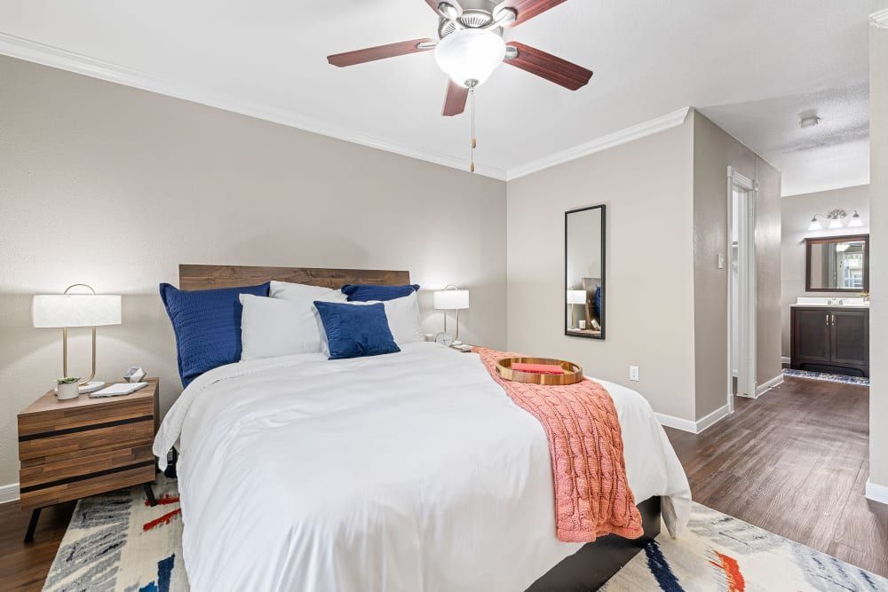 A modern bedroom with a large bed decorated with white and blue pillows and a coral throw blanket. There are two wooden bedside tables with white lamps, and a multi-colored area rug on the floor. A ceiling fan is mounted above, and the room opens to an en suite bathroom with a dark wood vanity visible in the background at Austin Midtown in North Austin, TX.