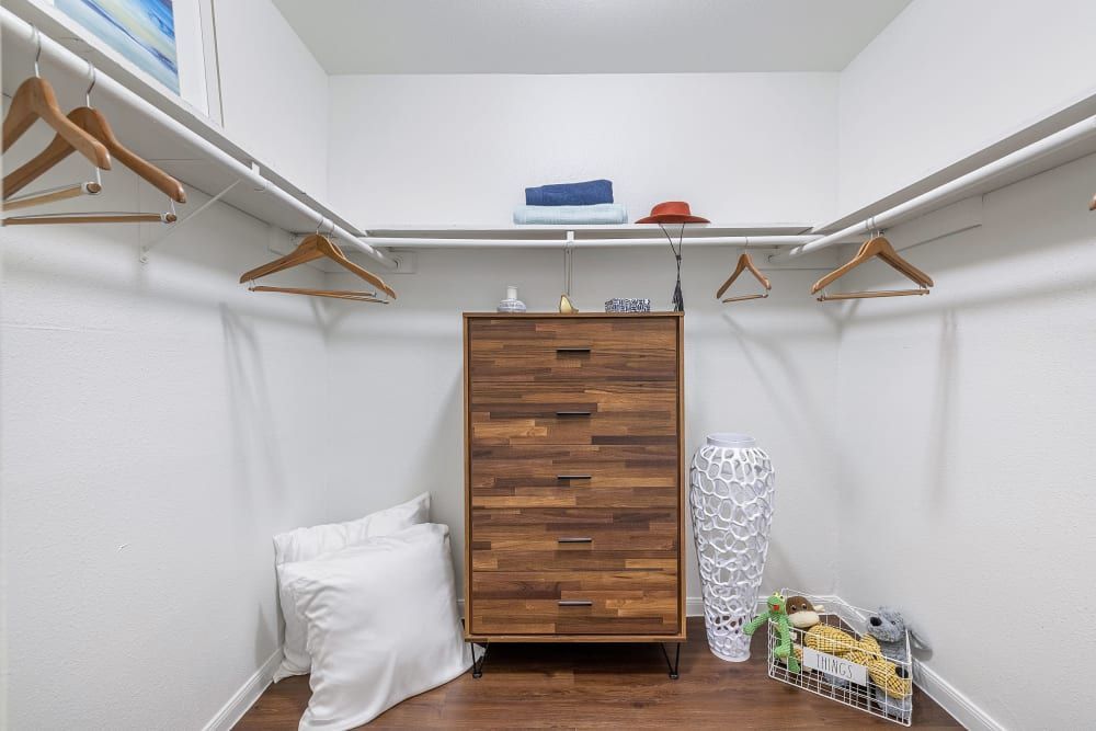 A tidy walk-in closet with white walls featuring a wooden dresser, a few empty hangers on closet rods, a decorative white vase, a pillow on the floor, folded towels on a shelf, and a wire basket with some items including a bottle and a bag on the floor at Austin Midtown in North Austin, TX.