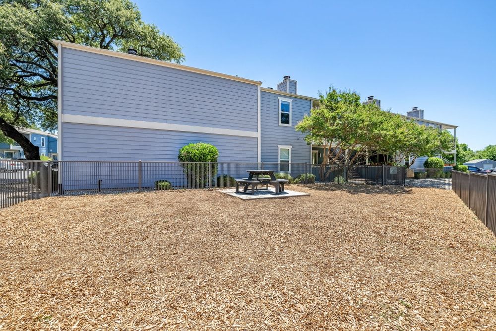  Exterior view of a modern gray townhouse with white trim, featuring a well-maintained dog park covered in wood chips, including a picnic table and surrounded by metal fencing with mature trees providing shade at Austin Midtown in North Austin, TX.