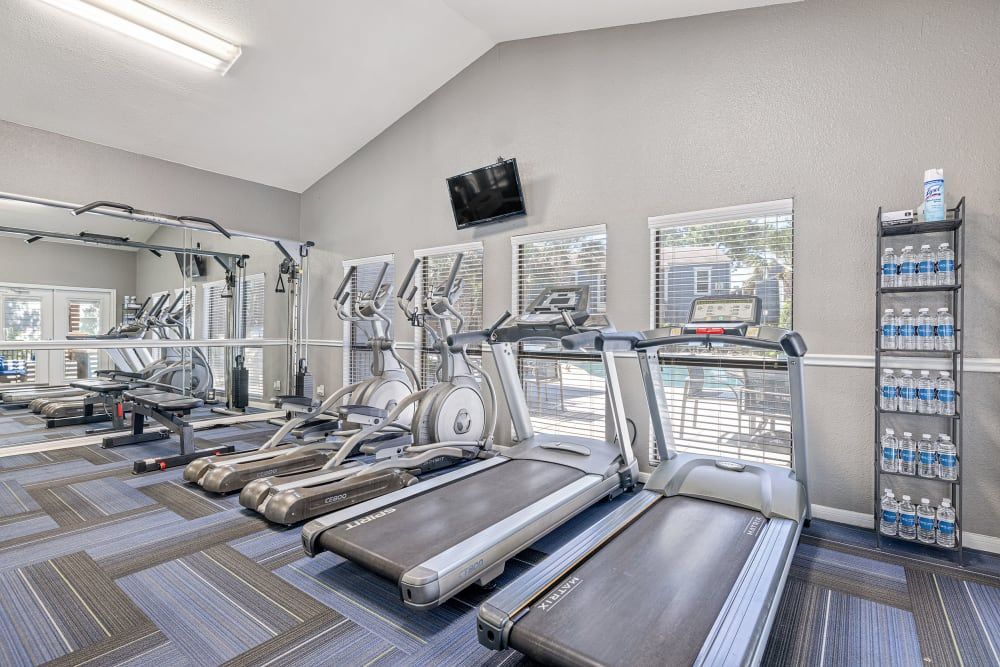 A well-lit, modern gym interior with a variety of exercise equipment including treadmills, elliptical machines, a weight bench, and a cable crossover machine. The room has gray walls, large windows with blinds, and a patterned carpet floor. A rack of bottled water is positioned to the side for hydration at Austin Midtown in North Austin, TX.