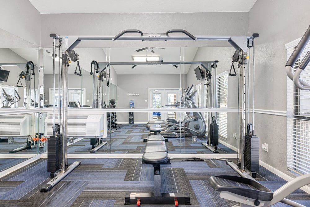 Modern gym interior with a variety of equipment including a multi-station cable machine, benches, dumbbells, and cardio machines on a patterned carpet floor, illuminated by natural light from windows with blinds at Austin Midtown in North Austin, TX.