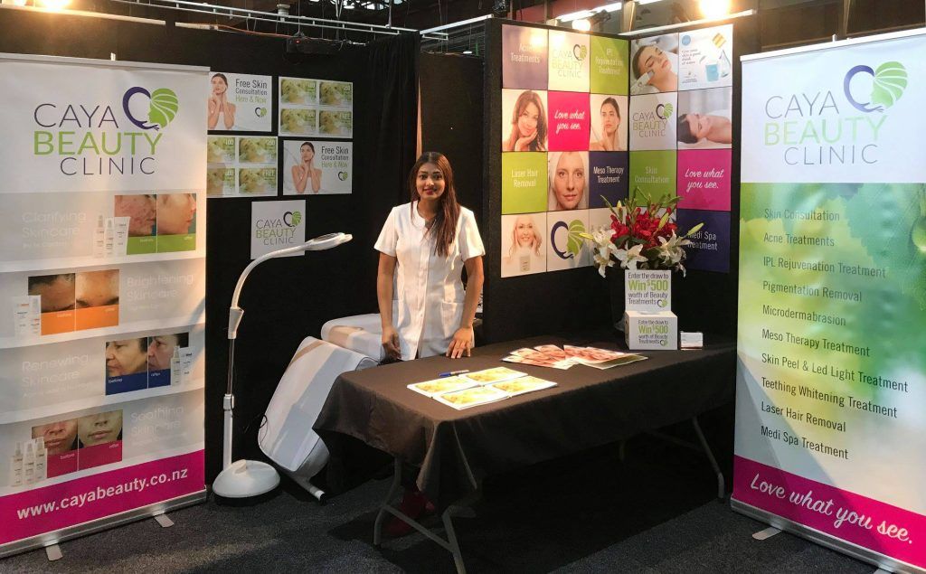 Trade show booth with beauty displays, a woman standing behind a black table, and colorful product posters.