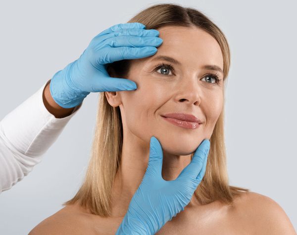 Gloved hands gently examine a woman’s face against a light gray background.