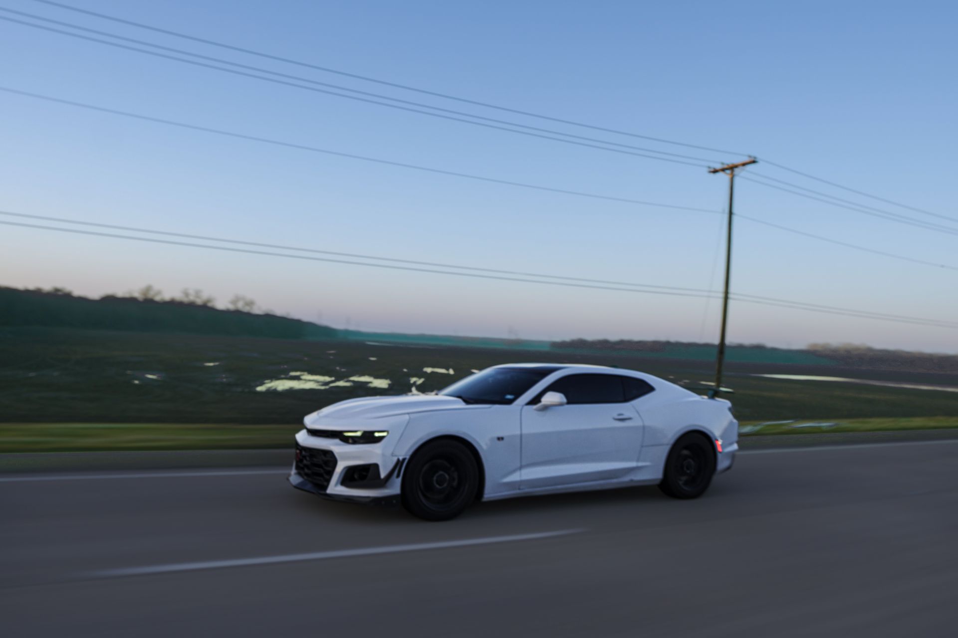 2019 Chevrolet Camaro 1LT in white and black in a rolling shot on the street in the morning, with greenery in the background.