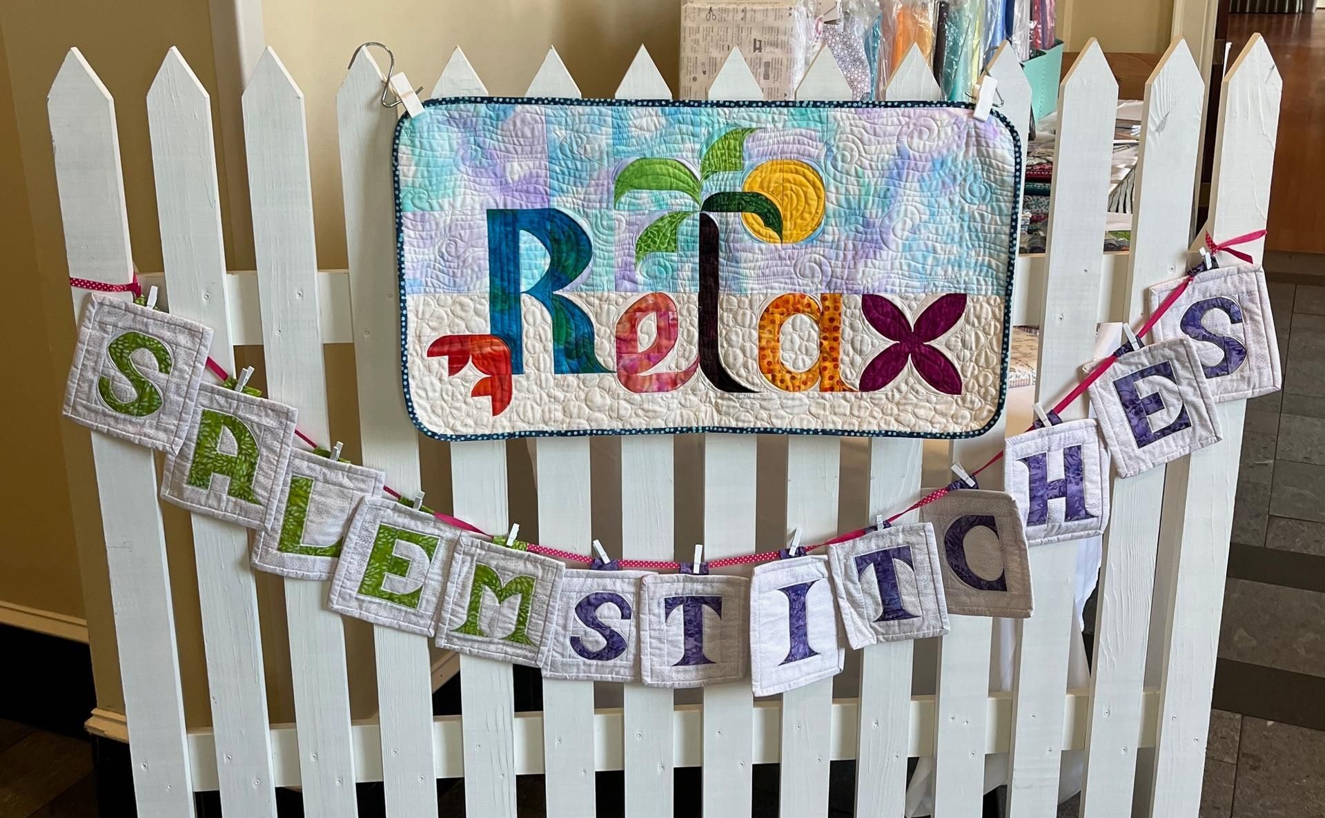 SaleM Stitches display booth with colorful fabrics, white picket fence, and reusable bag sign in a hallway