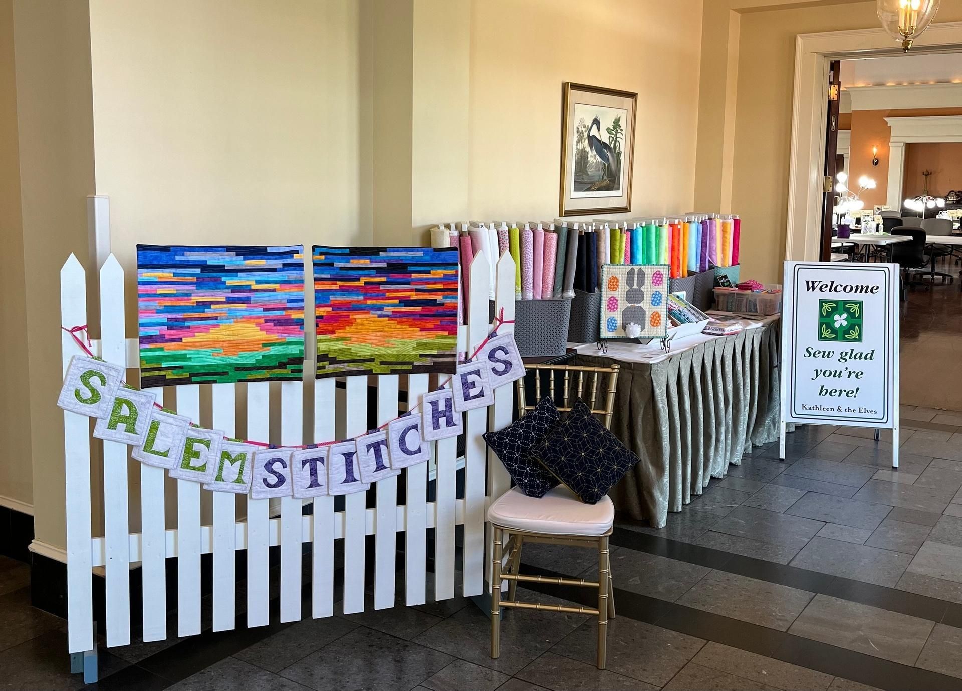 SaleM Stitches display booth with colorful fabrics, white picket fence, and reusable bag sign in a hallway