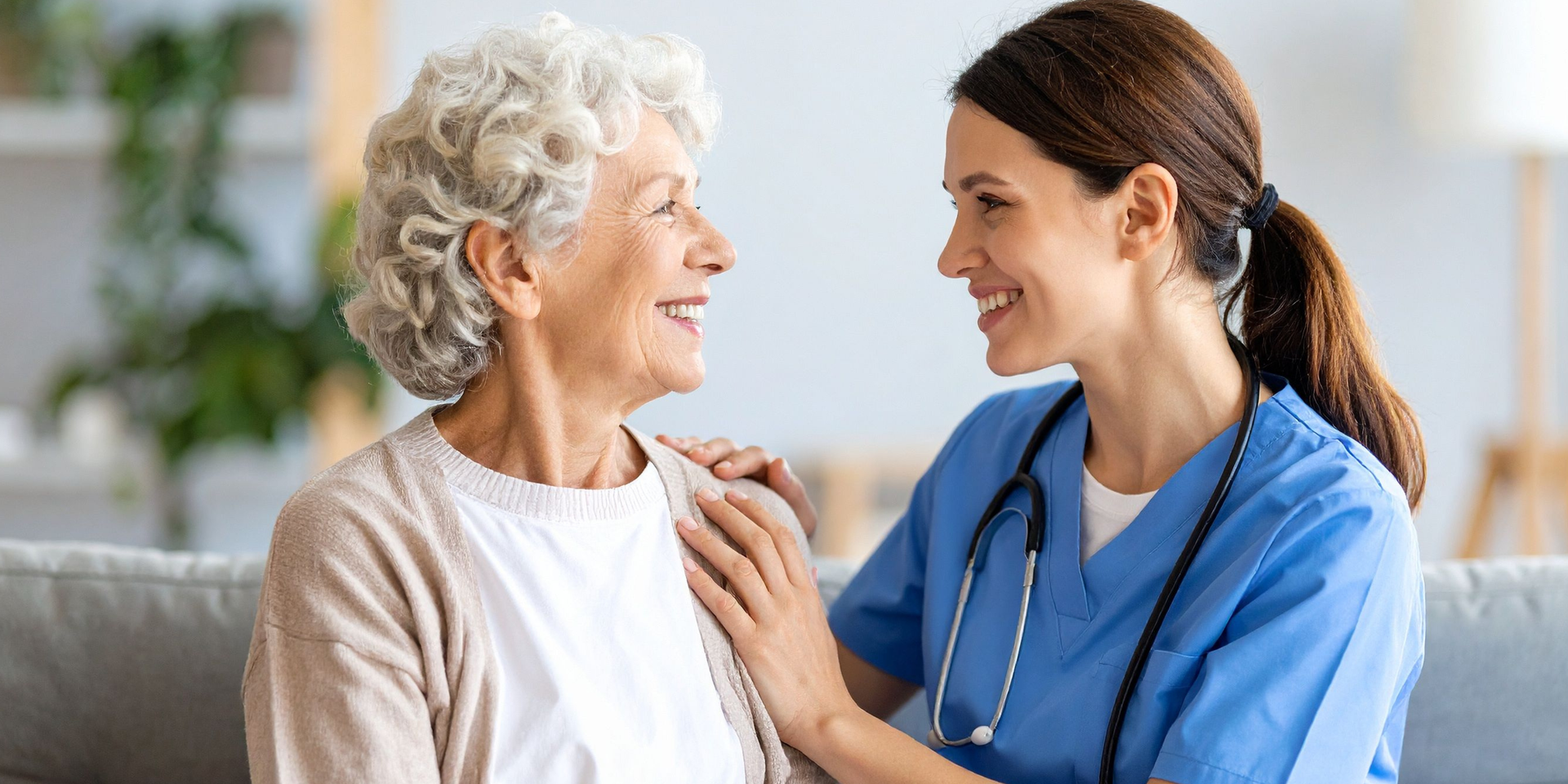 A healthcare provider in blue scrubs smiles at an older adult. The provider has an arm on the woman's shoulder; they are sitting on a couch.