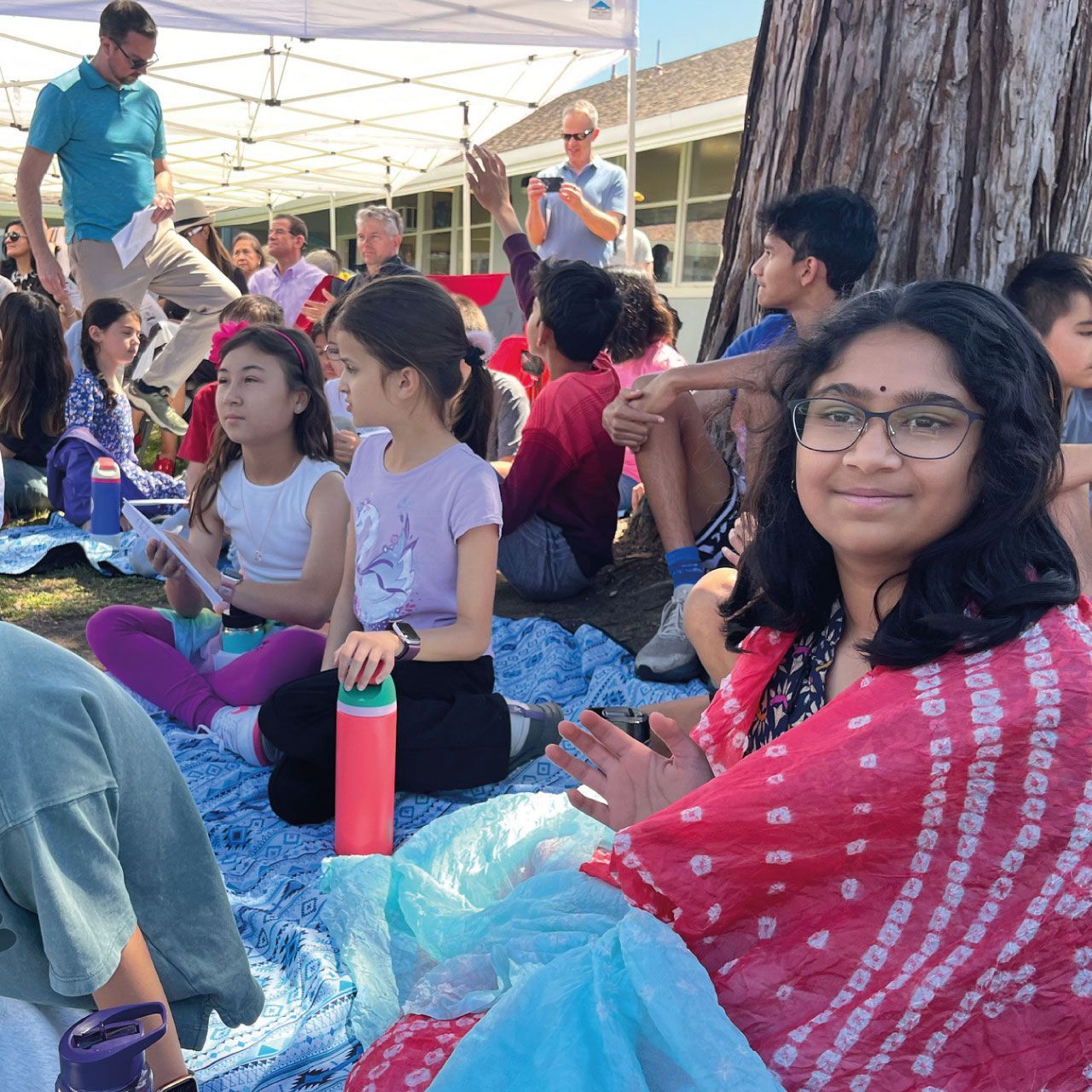 Montessori elementary students and families gather on picnic blankets outdoors under a canopy tent for a vibrant cultural celebration and school community event.