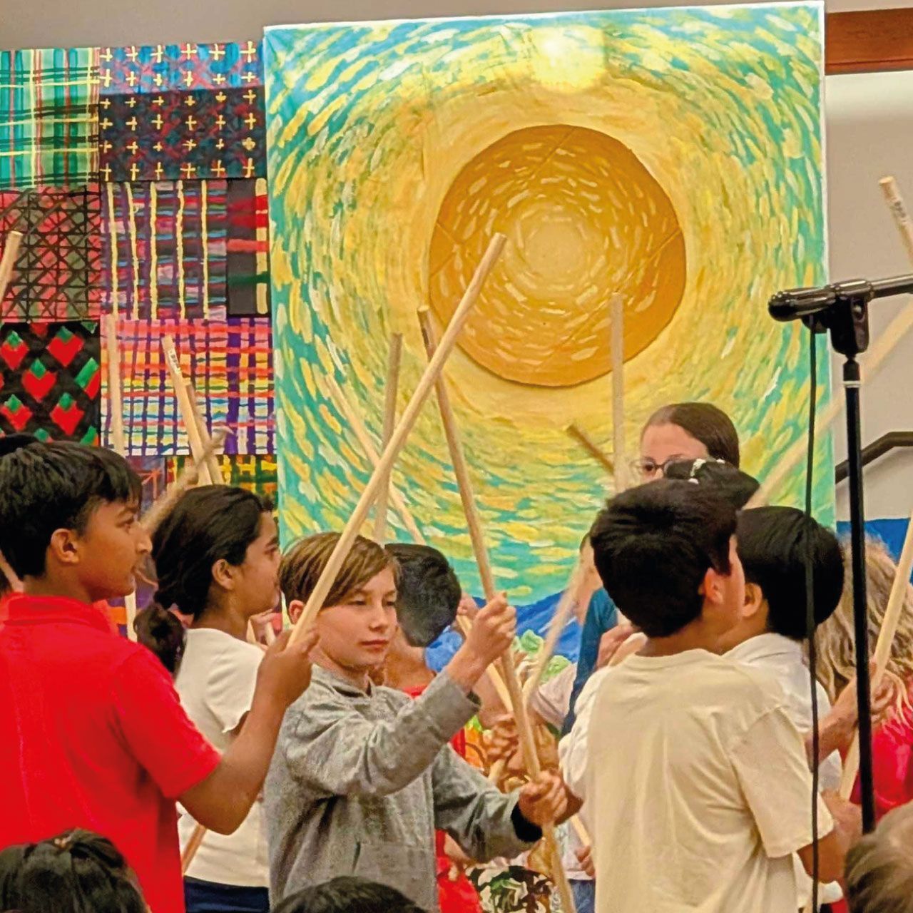 Montessori elementary students participate in a rhythmic stick performance on stage in front of a large, vibrant sun backdrop during a school arts showcase.