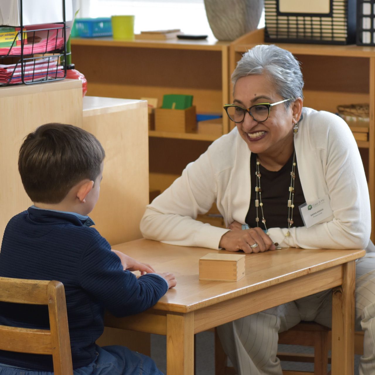 A Montessori primary guide smiles warmly at a young boy seated at a wooden table while giving a gentle one-on-one lesson in a prepared classroom environment.