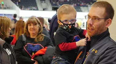 A man holds a child wearing a face mask at a stadium event, with several people standing in the background.