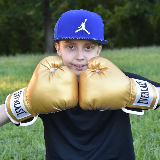 A person wearing a blue cap and gold Everlast boxing gloves holds their fists up in a fighting pose outdoors.