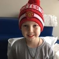 A smiling child wearing a red Manchester United beanie and a grey t-shirt, seated in front of a blue headboard.