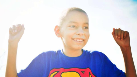 A person wearing a Superman t-shirt smiles with their arms raised in a bright, sunlit outdoor setting.
