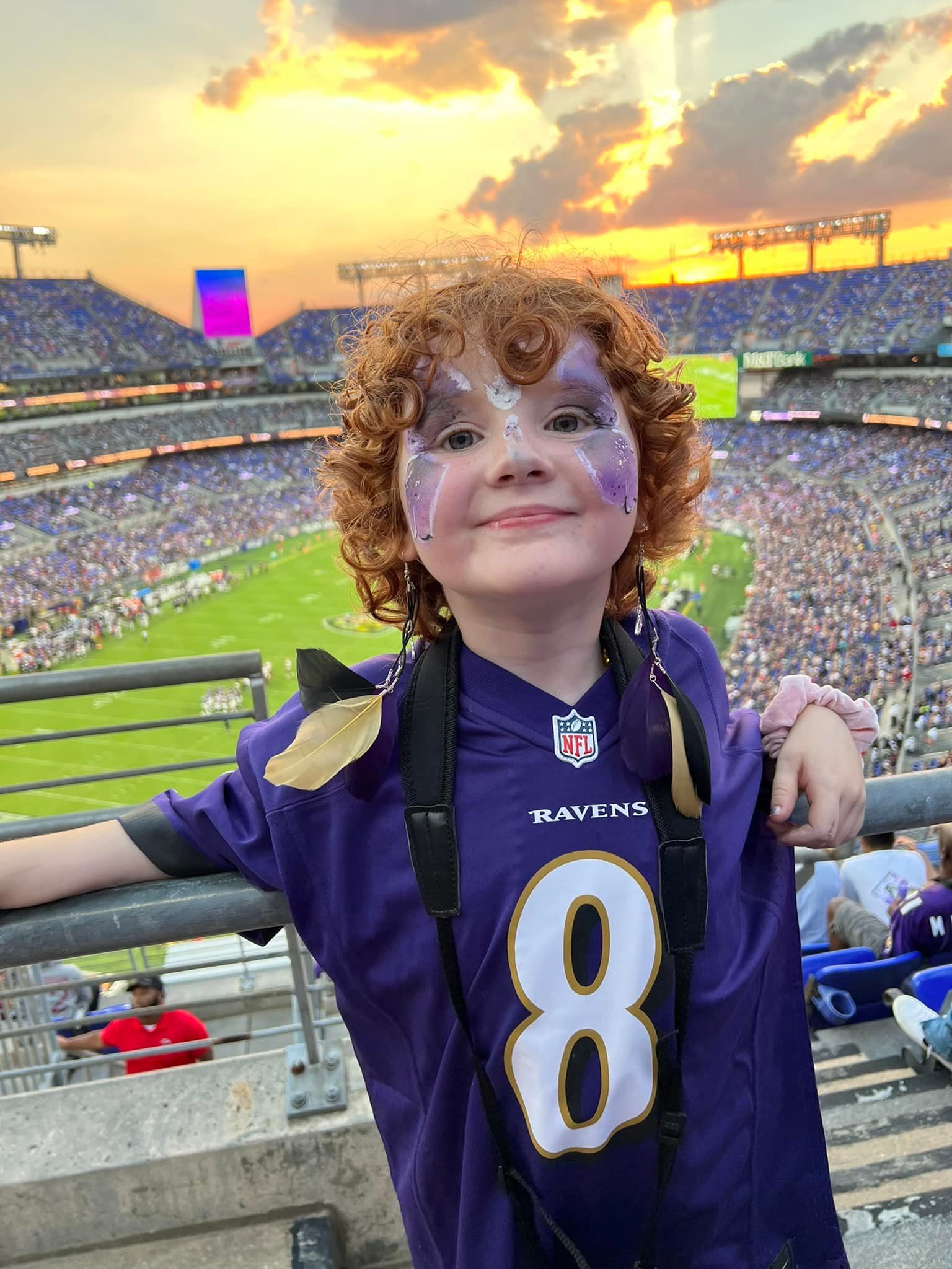 A person with curly hair and face paint wears a purple number 8 Baltimore Ravens jersey at a crowded football stadium.