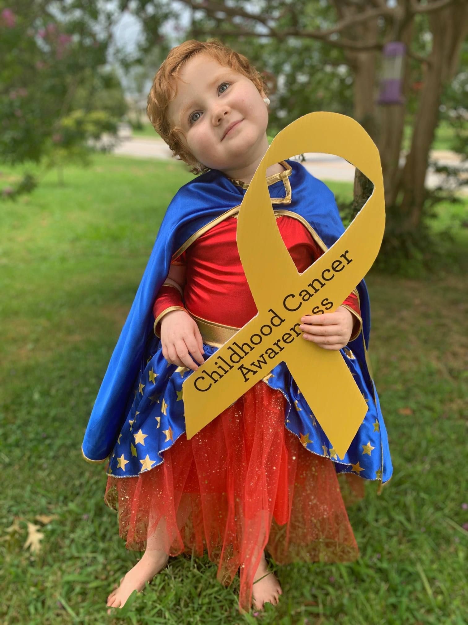 A child dressed as a superhero holds a yellow ribbon with the text 